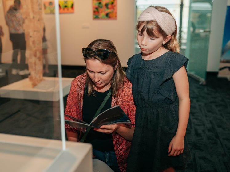 Image of A woman sitting and looking at a booklet whilst a young girl in a dark dress and headband stands beside her in an art gallery. Colourful artworks are displayed on the wall in the background.