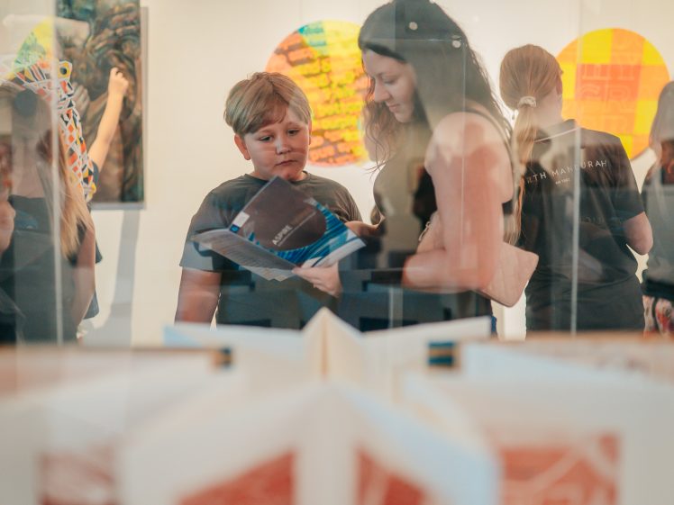 Image of A woman and a boy look at a book together in an art gallery. Other people and colourful artwork are visible in the background, with reflections and open books in the foreground.