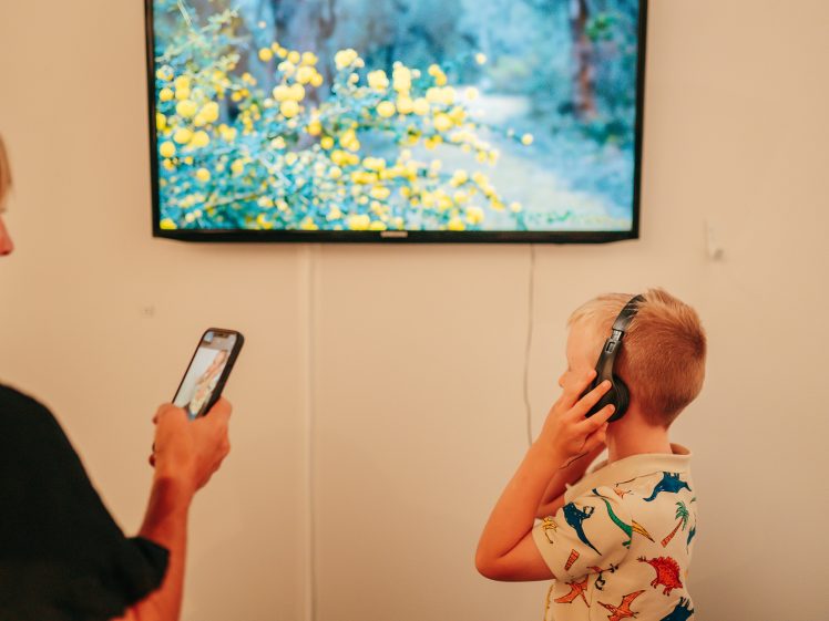 Image of A woman takes a photo with her phone while a young boy wearing headphones and a girl stand in front of a wall-mounted screen displaying yellow flowers.
