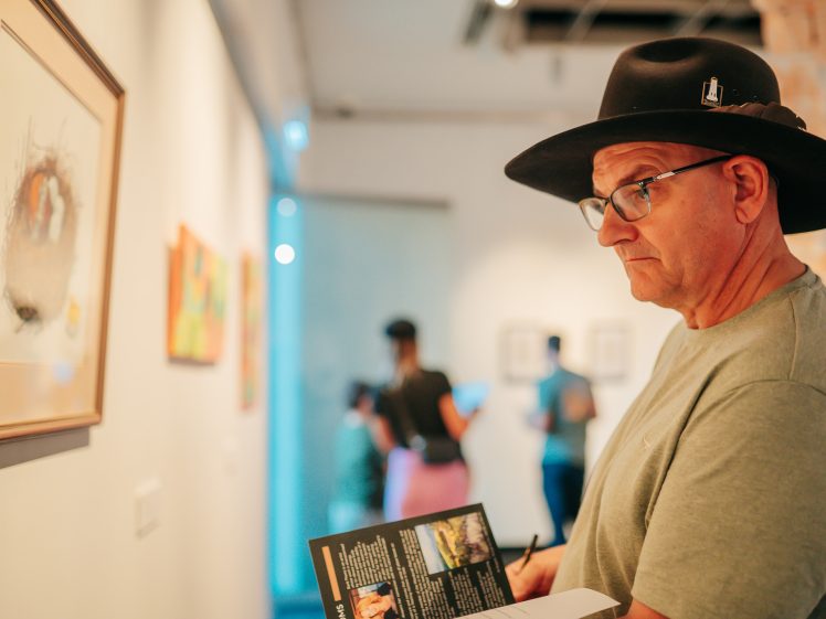 Image of A man in a black hat and glasses studies a framed artwork in a gallery, holding a pen and brochure. Blurred visitors and more art pieces are visible in the brightly lit background.