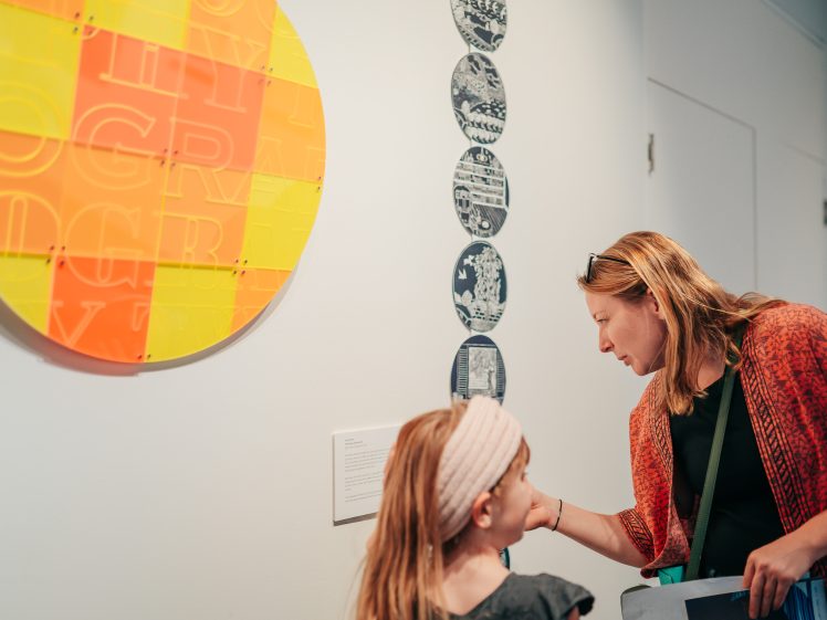 Image of A woman and a young girl look closely at art displayed on a gallery wall, including a large yellow-and-orange circular piece and a vertical row of smaller round artworks. The woman points at a label whilst speaking to the girl.