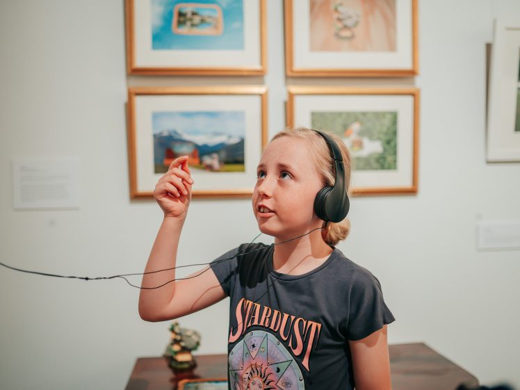 Image of A young girl wearing headphones and a Stardust T-shirt holds up an audio guide in an art gallery, looking up at framed artwork on the wall.