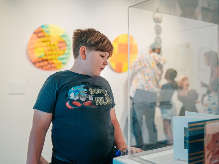Image of A boy with short brown hair and a dark graphic T-shirt looks at an exhibit in an art gallery, with colourful art pieces and other visitors visible in the background.