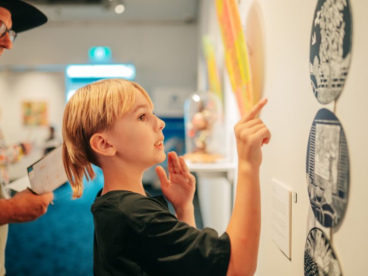 Image of A child points at artwork on a gallery wall while an adult stands nearby holding a piece of paper. The child looks engaged and curious, examining the displayed pieces closely. The setting is bright and modern.