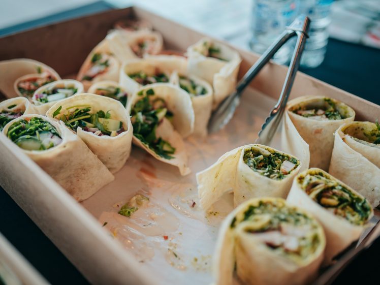 Image of A cardboard tray filled with assorted tortilla wrap sandwiches, cut into pieces and arranged in rows, with metal tongs resting on the tray. Some wraps contain salad leaves and other visible fillings.
