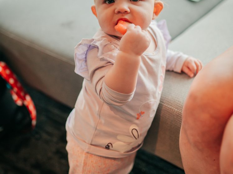 Image of A baby standing next to a sofa, holding and eating a snack with one hand, looking up towards the camera. The baby is wearing light-coloured clothes and has short blonde hair. An adult's legs are visible nearby.