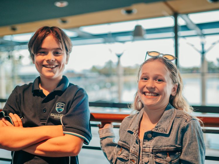 Image of Two smiling children stand indoors by a railing with large windows behind them; the boy wears a dark polo shirt, and the girl wears a denim jacket and sunglasses on her head. Natural light fills the scene.