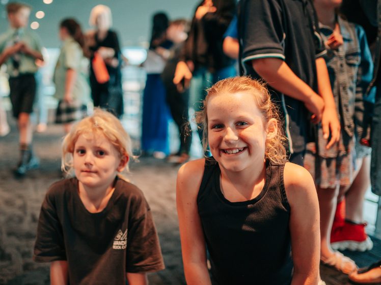 Image of Two young children are kneeling and smiling at the camera in the foreground, whilst a group of people and other children stand and talk in the blurred background indoors.