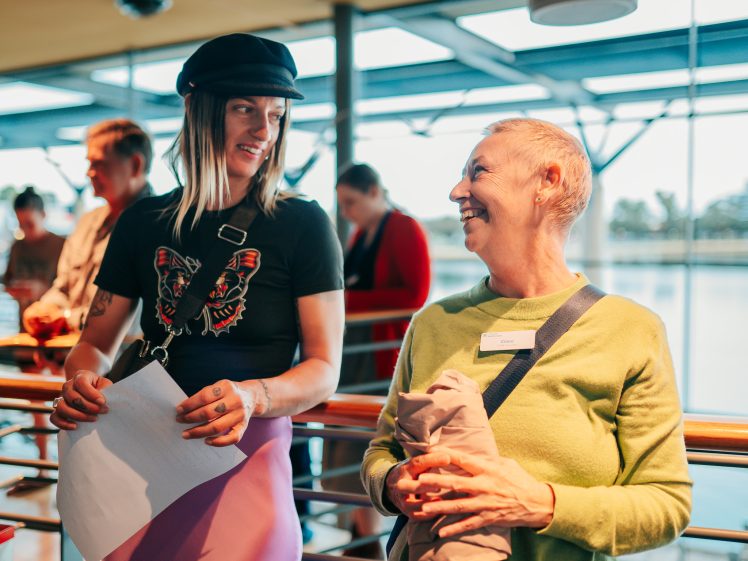Image of Two people stand indoors, smiling and talking. One wears a black hat and butterfly shirt, holding papers; the other wears a green jumper and holds a jacket, both wearing name badges. Others are blurred in the background.