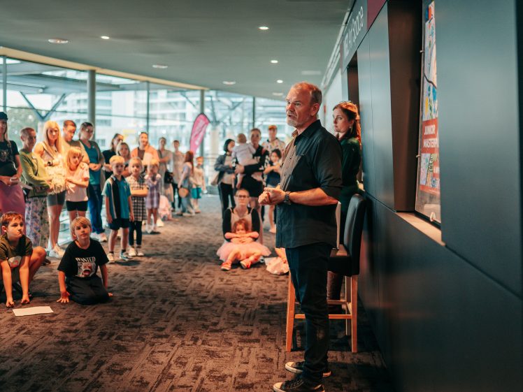 Image of A man stands speaking to a group of children and adults in a modern indoor space. Attendees listen attentively, some seated on the floor and others standing, with large windows and city views in the background.