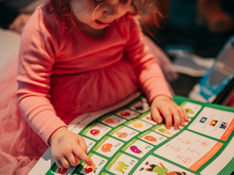Image of A young child in a pink dress sits on the floor, looking at and pointing to images and words on a colourful educational chart or poster.