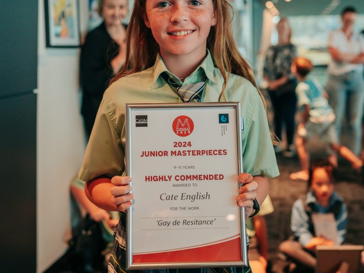 Image of A young girl in a green shirt and checked skirt smiles whilst holding a framed certificate for Highly Commended in the 2023 Junior Masterpieces art competition. Other people and artwork are visible in the background.