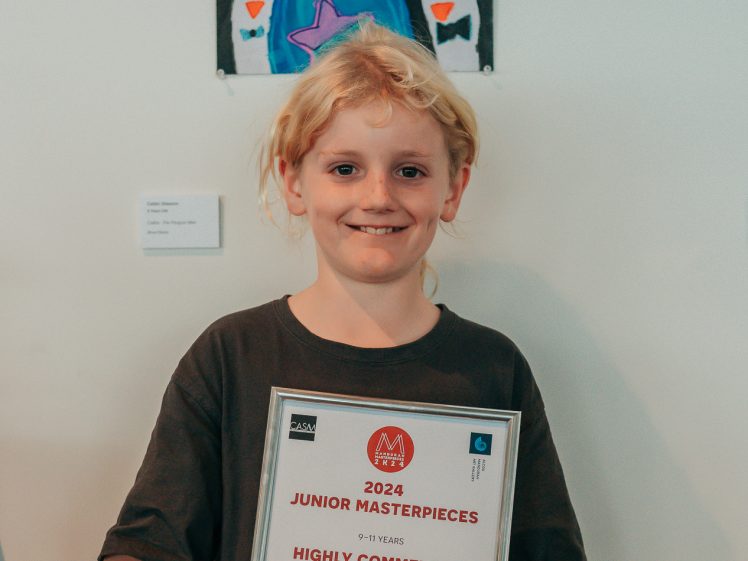 Image of A smiling child with blond hair holds a framed award certificate in front of a colourful penguin-themed painting. The certificate reads 2024 Junior Masterpieces, Highly Commended, Callan Glasson, Callan, The Penguin Man.