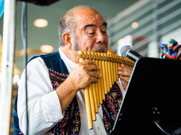 Image of An older man wearing a colourful, woven waistcoat plays a pan flute and sings into a microphone, performing indoors with music sheets in front of him.