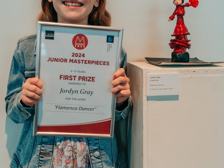 Image of A smiling girl holds a framed certificate for winning first prize in the 2024 Junior Masterpieces art competition. Beside her is a small sculpture titled Flamenco Dancer displayed on a white plinth.