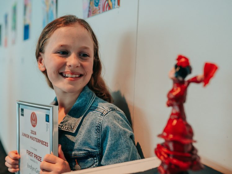 Image of A smiling girl in a denim jacket holds a certificate whilst admiring a small flamenco dancer figurine in a gallery with colourful artwork on the wall behind her.