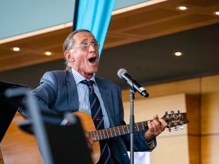 Image of An older man in a suit sings passionately into a microphone whilst playing an acoustic guitar indoors, under warm ceiling lights.