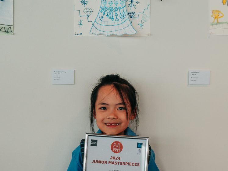 Image of A young girl smiles while holding a framed certificate for first prize in a junior art competition, standing in front of her crayon drawing of a girl with plaited hair and a blue dress.