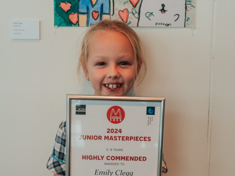 Image of A smiling young girl in a chequered dress holds a certificate reading 2024 Junior Masterpieces Highly Commended for her artwork Pushen and Me. Behind her is her colourful drawing of a girl and a cat with hearts.