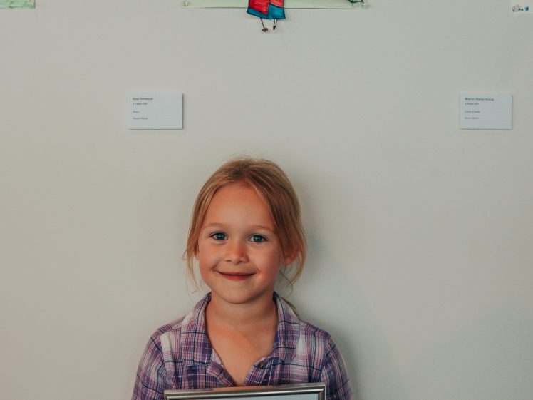 Image of A young girl in checked pyjamas smiles whilst holding a framed certificate in front of her colourful artwork displayed on a wall. The certificate reads Highly Commended for Junior Masterpieces 2024.