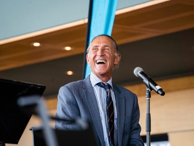 Image of A man in a suit and striped tie smiles and laughs whilst standing at a microphone indoors, with modern lighting and a blue banner in the background.