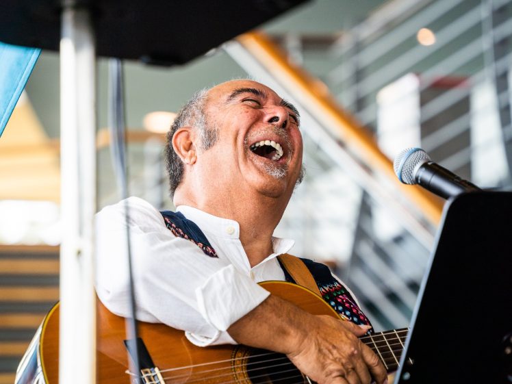 Image of A man joyfully plays an acoustic guitar and sings into a microphone, laughing with his eyes closed. He wears a white shirt and sits indoors near a staircase.