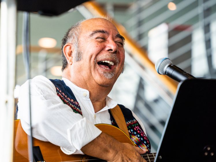Image of A smiling man with a guitar sits behind a microphone, laughing joyfully. He wears a colourful waistcoat over a white shirt, and the background shows a modern interior with stairs and railings.