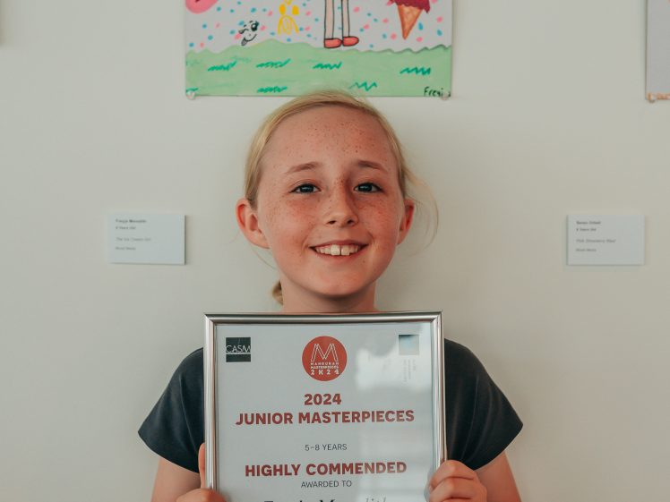 Image of A smiling young girl holds a framed certificate for a Highly Commended junior art award, standing in front of her colourful drawing of a girl, heart, and star. She wears a dark T-shirt with a magical theme.
