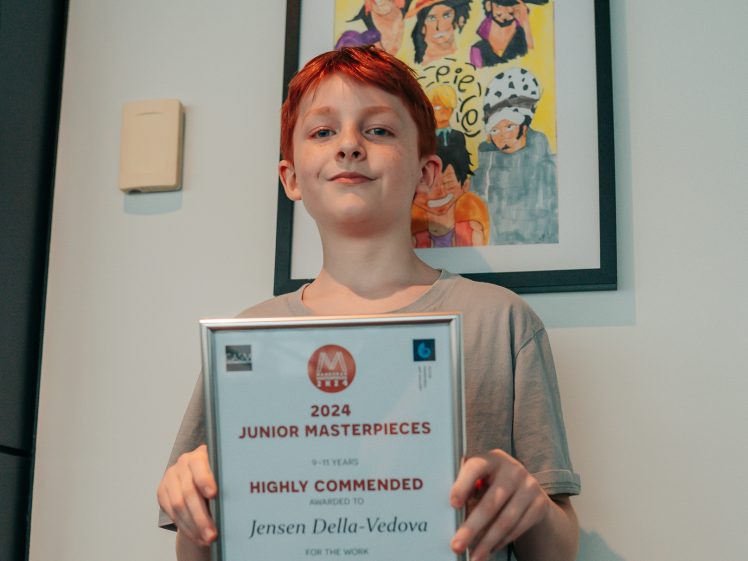 Image of A young person with red hair holds a framed certificate in front of a colourful painting. The certificate reads 2024 Junior Masterpieces Highly Commended awarded to Jensen Della-Vedova for the work Friends.
