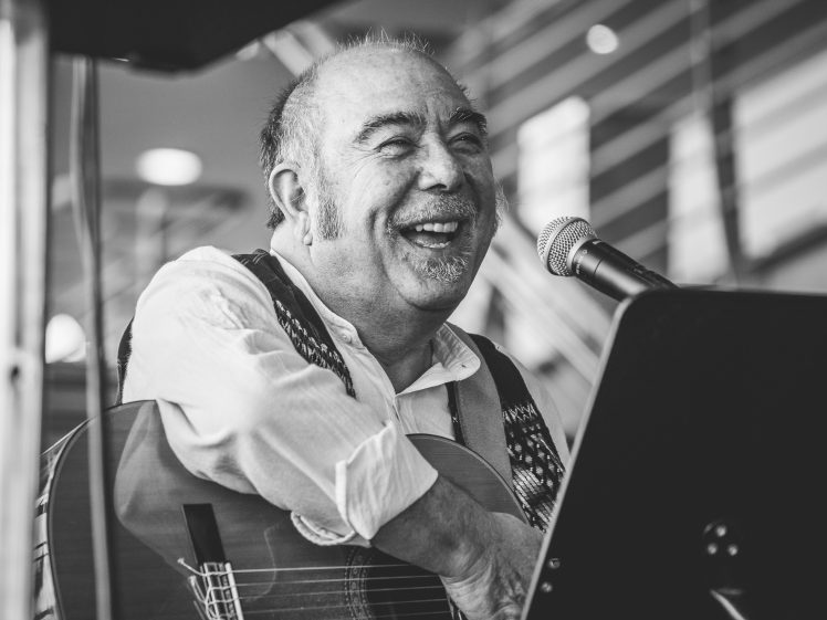Image of A smiling older man with sideburns plays an acoustic guitar and sings into a microphone. He is wearing a patterned waistcoat over a light shirt. The image is black and white, and he appears to be performing indoors.