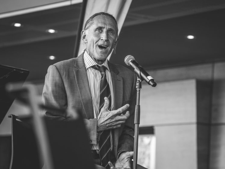 Image of An older man in a suit sits and sings or speaks passionately into a microphone. The image is in black and white, and the background shows an indoor modern setting with lights and architectural details.