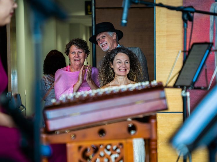 Image of Three smiling adults, two women and one man in a hat, stand closely together indoors, applauding. Musical instruments and equipment are visible in the foreground, suggesting a musical performance or event.