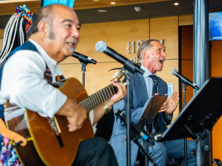 Image of A man in a suit sings into a microphone whilst clapping, next to another man playing guitar and singing, both performing indoors on a brightly lit stage.