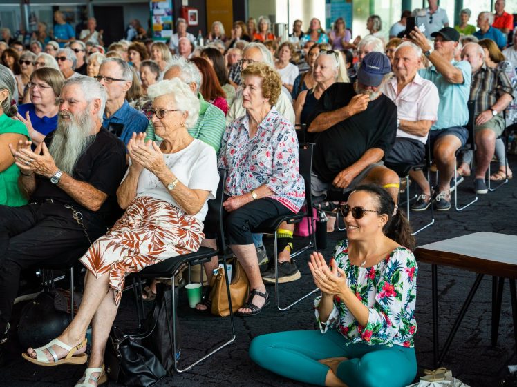 Image of A large group of people, mostly older adults, sit and clap at an indoor event. A woman in colourful clothing sits cross-legged on the floor in front, also applauding, while others sit in chairs behind her.