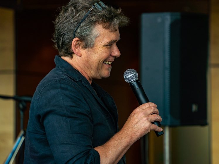 Image of A man with grey hair, wearing a black shirt and glasses on his head, smiles while speaking into a microphone indoors. A large speaker and blurred background are visible behind him.