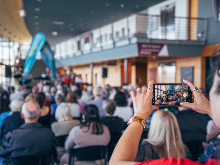 Image of A person holding a mobile phone takes a photo of a seated audience at an indoor event, with a stage and people visible in the background. The image is focused on the phone's screen displaying the scene.