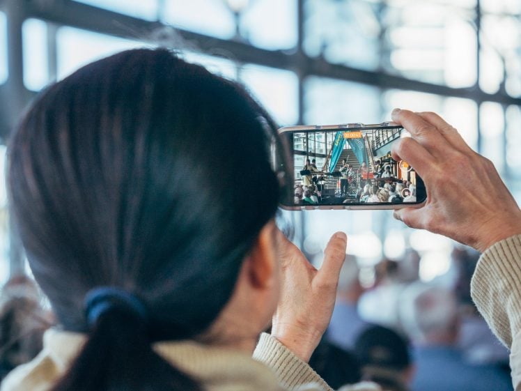 Image of A person with dark hair takes a photo or video of a public event using a mobile phone, capturing a crowd and a performer on stage in a bright, spacious venue with large windows.
