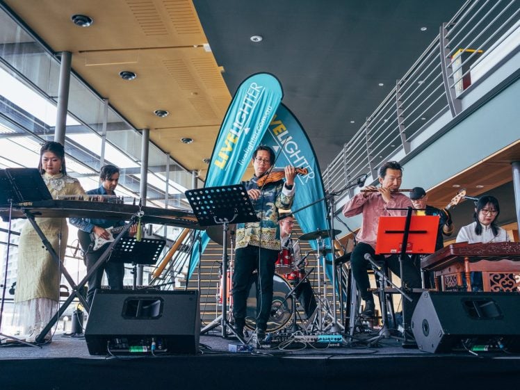Image of A group of musicians play traditional and modern instruments on a stage indoors; a blue Live Lighter banner stands behind them, and sunlight streams through large windows.