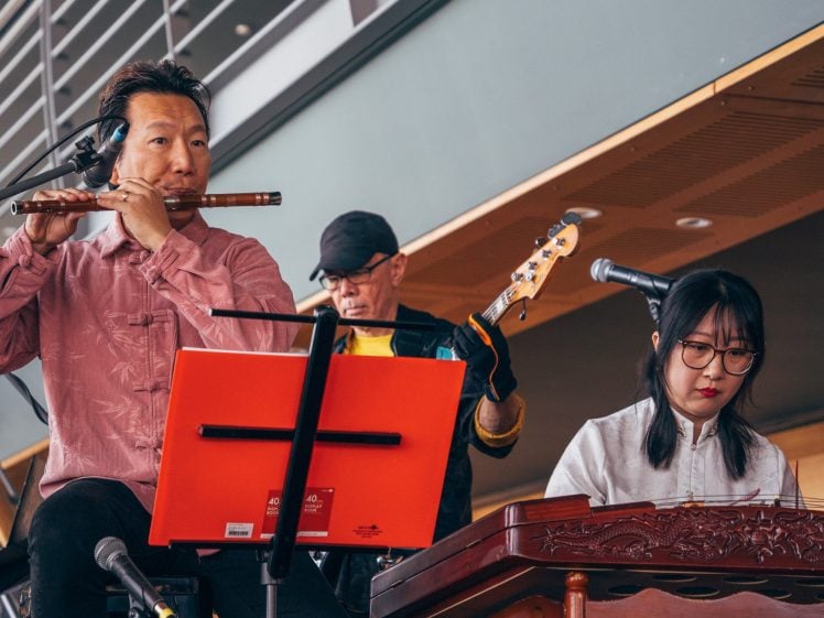 Image of A man plays the flute, another plays electric bass, and a woman plays a traditional string instrument at an indoor music performance. Sheet music is set up in front of them.