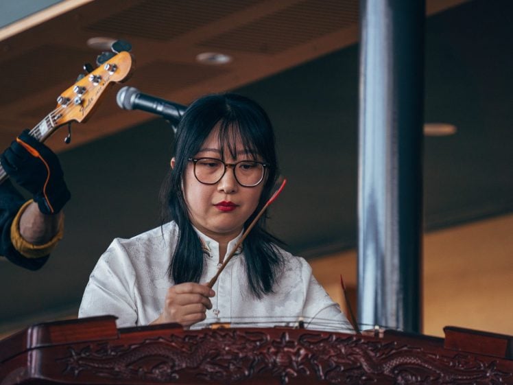 Image of A woman in glasses and a white traditional top plays a stringed instrument, concentrating intently. A microphone and part of a guitar are visible in the background.