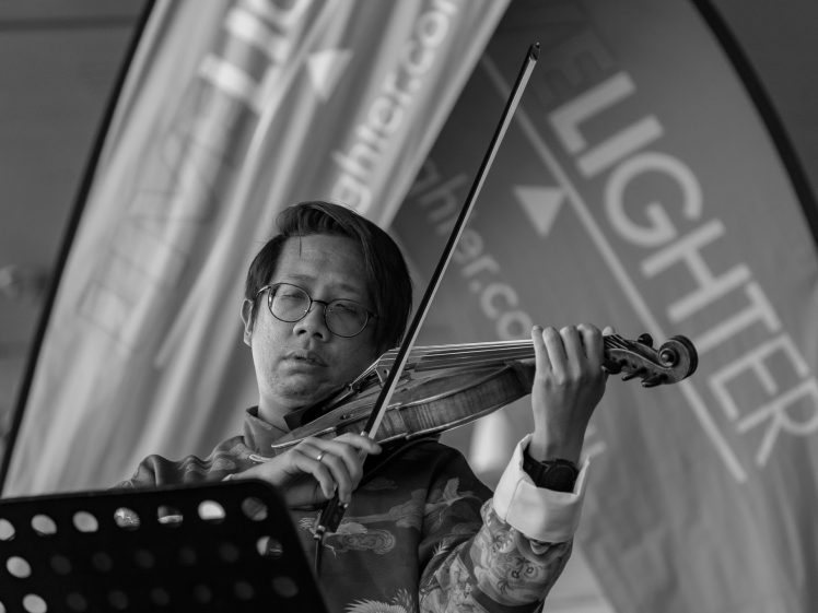 Image of A man wearing glasses plays a violin, focused on his performance. Behind him are blurred banners with partially visible text. The image is in black and white.