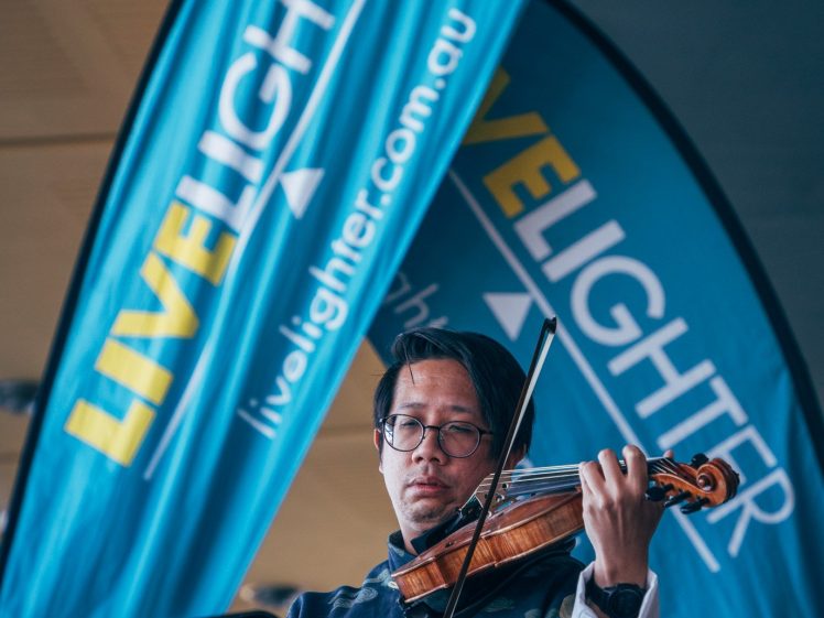 Image of A musician wearing glasses and a patterned jacket plays the violin in front of two large blue LiveLighter promotional banners at an indoor event.