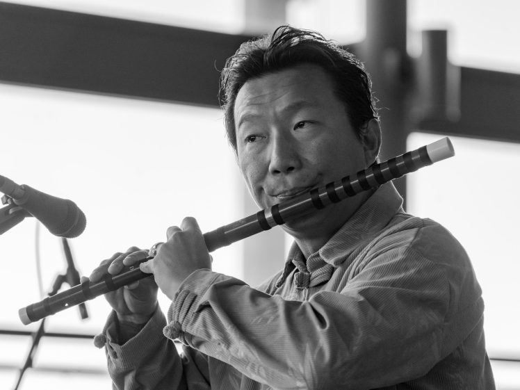 Image of A person playing a flute near a microphone, wearing a collared shirt. The image is in black and white with soft lighting and a blurred background.