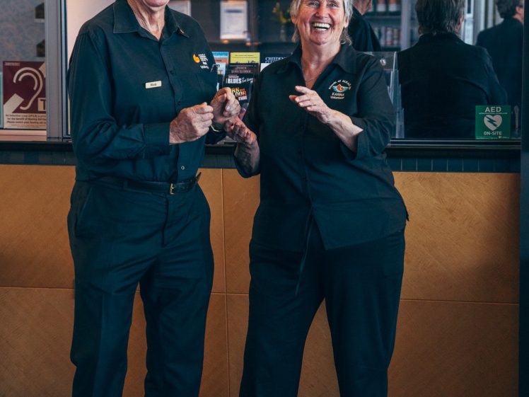 Image of Two older adults in matching black uniforms stand and laugh together in front of a counter, holding a leaflet. There is a mirror behind them reflecting their backs and part of the room.