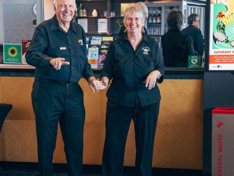 Image of Two older adults in matching black uniforms smile and gesture while standing side by side in a modern indoor setting near a counter with leaflets and posters.