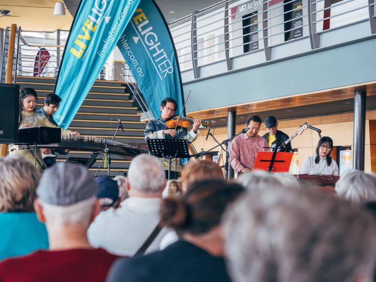 Image of A band performs on stage with various instruments, including a violin, keyboard, and guitar, in front of a seated audience indoors. Blue LiveLighter banners and a staircase are visible in the background.