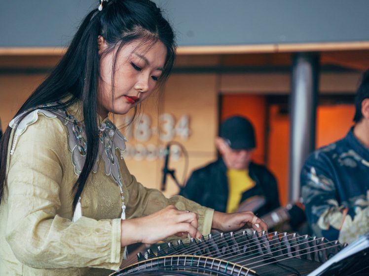 Image of A woman with long black hair plays a traditional stringed instrument, focused on her performance, whilst musicians in the background play guitars.