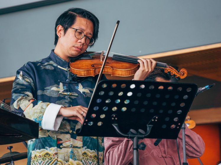 Image of A musician wearing glasses and a patterned traditional-style jacket plays the violin while reading sheet music from a stand during a performance.