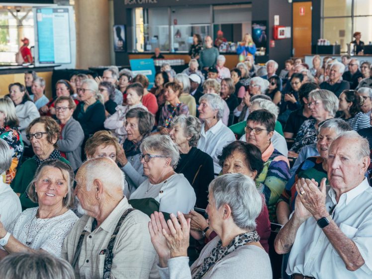 Image of A large group of elderly people sit closely together indoors, some applauding and smiling, as they watch an event or performance. The atmosphere appears lively and engaged.