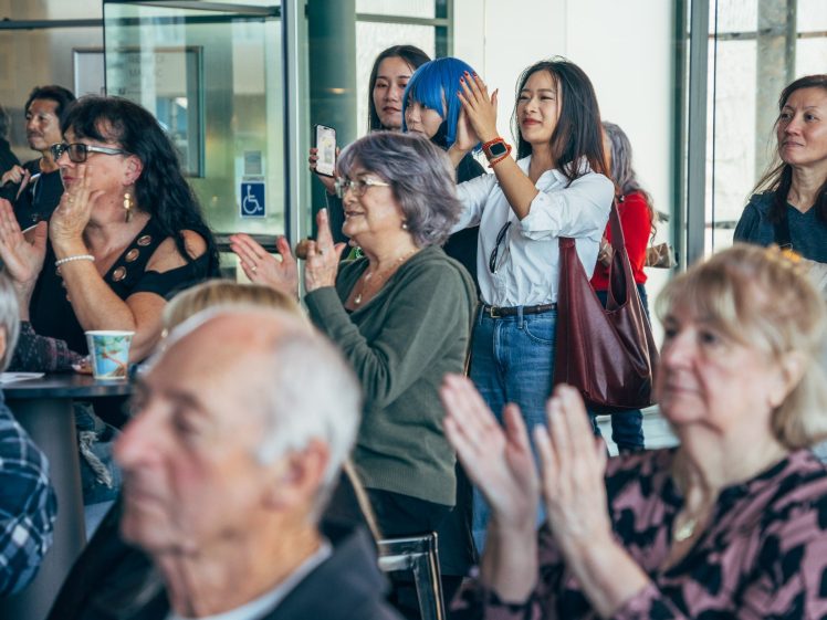 Image of A diverse group of people sit and stand indoors, clapping and smiling whilst watching an event. Some are taking photos with their mobiles, and the atmosphere appears lively and engaged.